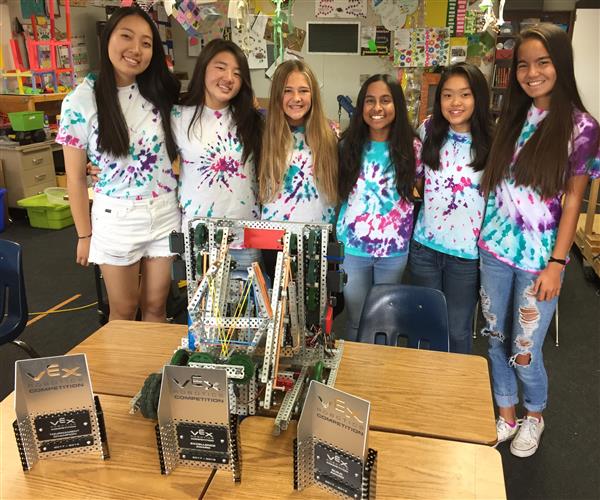 Photo of six high school girls in tye-dye shirts, standing behind a robot they built